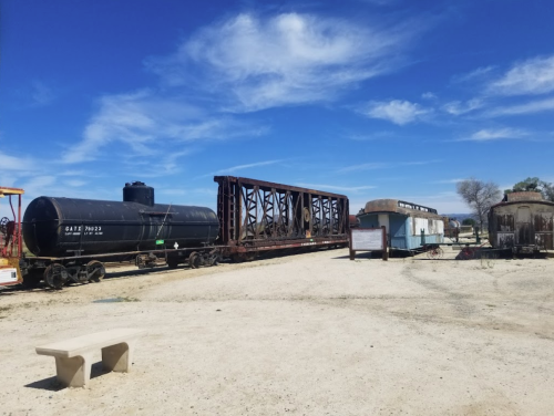 A train car and a flatbed on a dusty landscape under a blue sky, with a few structures in the background.