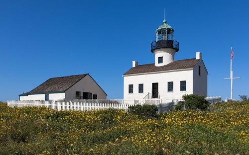 A white lighthouse with a green dome, surrounded by yellow flowers and a white picket fence under a clear blue sky.