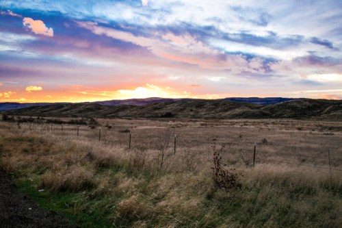 A scenic landscape at sunset, featuring rolling hills, a colorful sky, and a grassy field with a fence in the foreground.