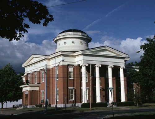 Historic building with a dome and columns, set against a blue sky with clouds. Green trees in the foreground.