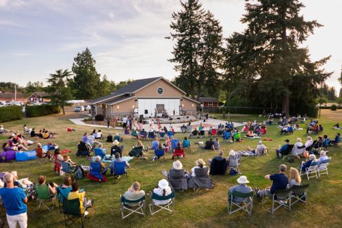 A large crowd sits on lawn chairs and blankets, enjoying a live performance at an outdoor venue surrounded by trees.