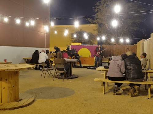 A cozy outdoor venue with string lights, featuring a live band and people enjoying the music at wooden tables.