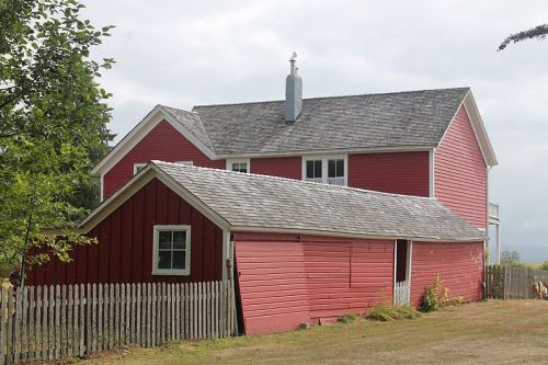 A red farmhouse with a slanted roof and a wooden fence, surrounded by greenery under a cloudy sky.