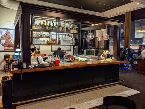 A stylish bar with shelves of liquor, a bartender, and patrons in a modern restaurant setting.