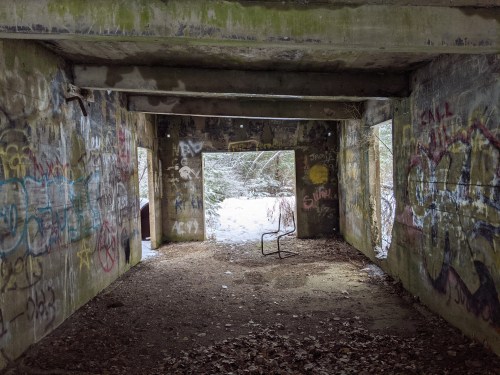 Abandoned room with graffiti-covered walls, two open windows, and a lone chair on a dirt floor. Snow visible outside.