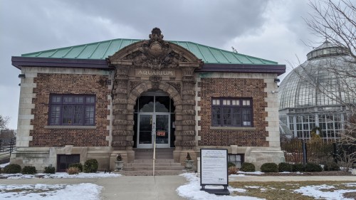 Historic aquarium building with ornate entrance, green roof, and a glass conservatory nearby, set against a cloudy sky.
