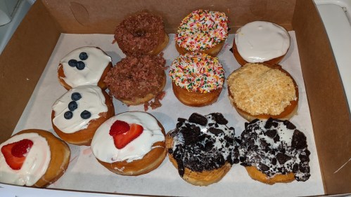 A box of assorted donuts topped with frosting, sprinkles, fruit, and cookie crumbles in various flavors.