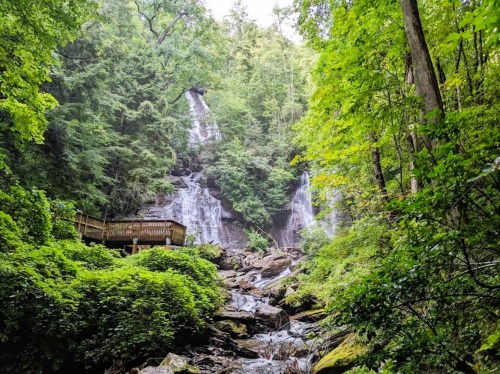 A serene waterfall cascades down rocky terrain, surrounded by lush green trees and foliage. A wooden platform is visible nearby.