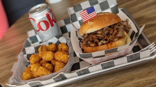 A tray with a pulled pork sandwich topped with a small American flag, a side of tater tots, and a can of Coke.