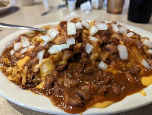 A plate of chili cheese fries topped with chopped onions and beans, served with a fork on a table.