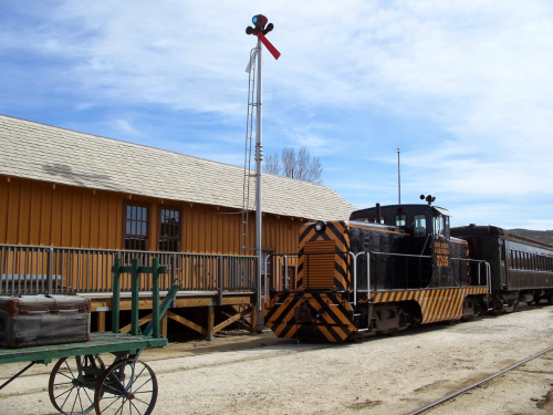 A vintage train engine parked beside a yellow building, with a signal tower and a green cart in the foreground.