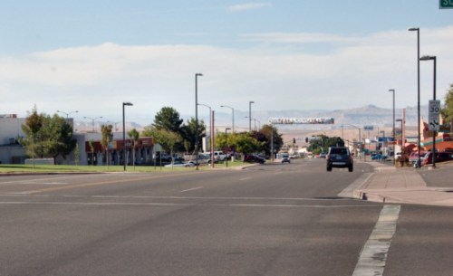 A wide street view with shops on the left, cars parked, and distant mountains under a clear blue sky.