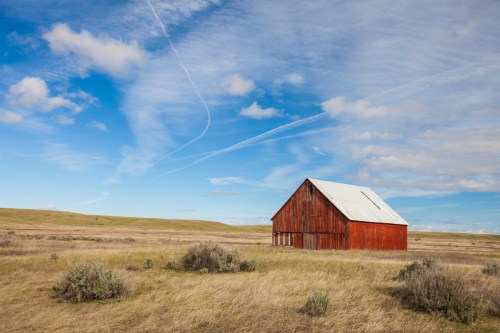 A red barn stands alone in a grassy field under a blue sky with wispy clouds and distant hills.