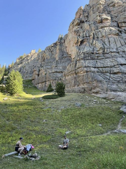 Two people sit on a grassy area near a rocky cliff, surrounded by trees and clear blue skies.