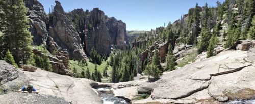 A panoramic view of a rocky canyon with lush green trees and a small waterfall in the foreground.