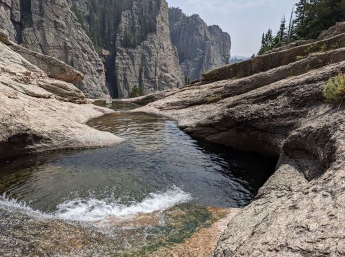 A serene rocky landscape with a clear pool of water surrounded by towering cliffs and trees in the background.