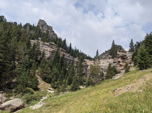 Rocky cliffs rise above a lush green hillside, surrounded by dense pine trees under a partly cloudy sky.