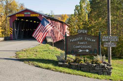 A red covered bridge with an American flag, leading to Paradise Stream Family Campground and Mini-Golf entrance.