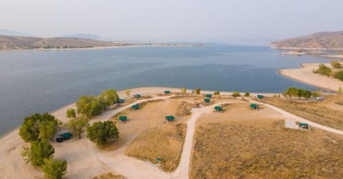 Aerial view of a lakeside campsite with green tents, surrounded by sandy shores and gentle hills in the background.