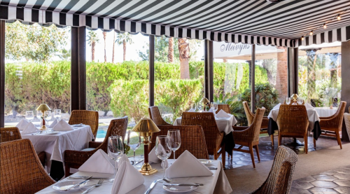 Elegant restaurant interior with striped awning, white tablecloths, and wicker chairs, surrounded by greenery.