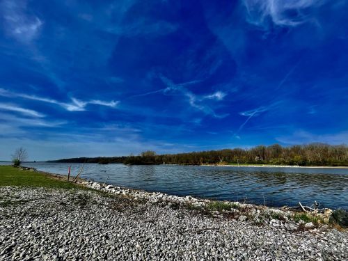 A serene river scene with a clear blue sky, lush green banks, and rocky shoreline under a bright, sunny day.
