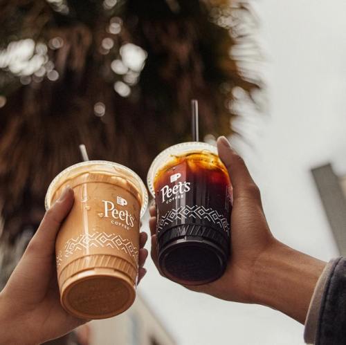 Two hands holding Peet's Coffee cups, one iced coffee and one iced latte, against a backdrop of palm trees.
