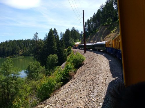 A train curves along a scenic route by a lake, surrounded by lush trees and a clear blue sky.