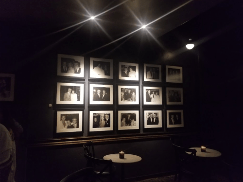 A dimly lit wall featuring framed black-and-white photographs, with small tables and candles in a cozy setting.