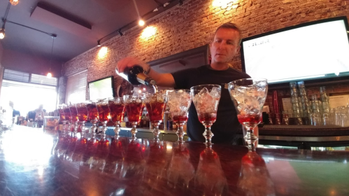 A bartender pours a drink into a row of glasses filled with ice at a lively bar with brick walls.