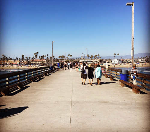 A sunny pier with people walking, lined with palm trees and ocean views in the background.