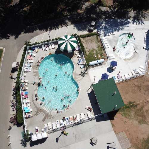 Aerial view of a pool area with two pools, lounge chairs, and people enjoying the sun surrounded by greenery.