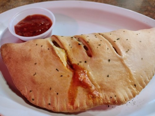 A golden-brown calzone with slits on top, served with a small cup of marinara sauce on a white plate.