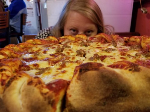 A person peeks over a large, cheesy pizza with stuffed crust, showcasing toppings and a vibrant restaurant background.