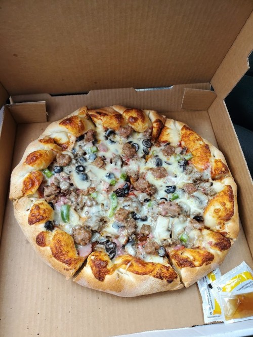 A pizza in a cardboard box topped with cheese, meats, olives, and green peppers, with a side of dipping sauce.