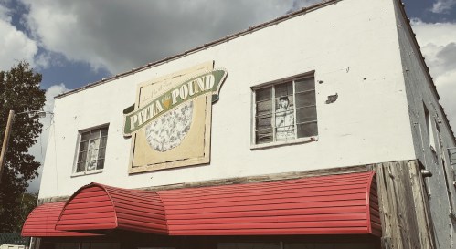 A building with a red awning and a sign reading "Pizza A Pound" under a cloudy sky. Windows are visible above the entrance.