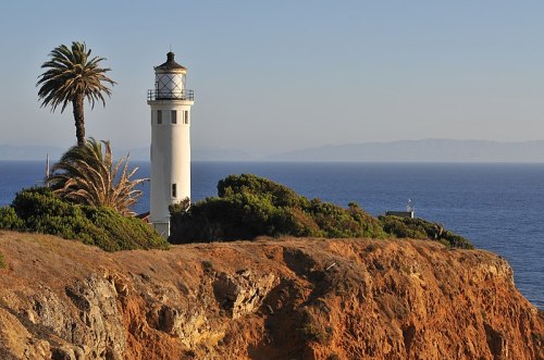 A white lighthouse stands on a rocky cliff, surrounded by greenery and palm trees, overlooking the ocean.