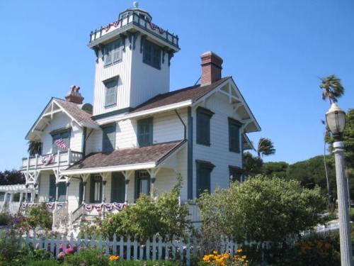 A charming white house with a tower, surrounded by greenery and flowers, featuring decorative elements and a picket fence.