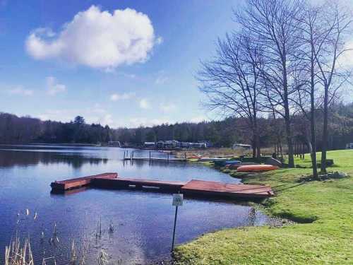 A serene lake scene with a wooden dock, kayaks, and trees under a blue sky with fluffy clouds.