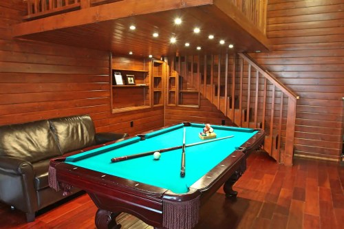 A cozy wooden room featuring a pool table, leather couch, and a staircase in the background.