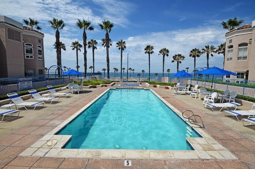 A clear swimming pool surrounded by lounge chairs and palm trees, with a beach view in the background.