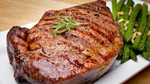 A grilled steak garnished with rosemary, served alongside asparagus and pine nuts on a white plate.