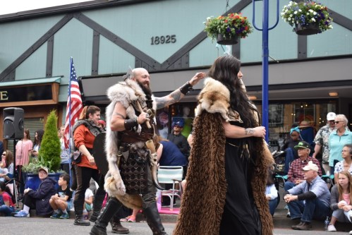 A parade scene featuring two individuals in fur costumes, with onlookers and an American flag in the background.
