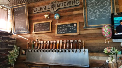 A wooden bar with multiple beer taps and a chalkboard menu at Valhöll Brewing, featuring various beer options.
