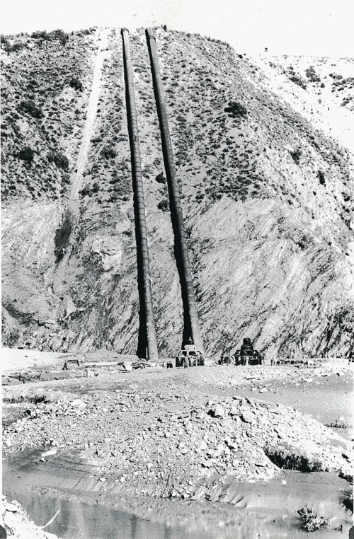 Black and white photo of two large pipes on a hillside, with machinery and rocky terrain in the foreground.