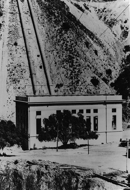 Historic black and white photo of a building at the base of a mountain, with pipelines running down the slope.