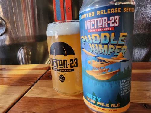 A can and glass of Victor-23 Puddle Jumper Hazy IPA on a wooden table, showcasing vibrant branding and a golden beer.