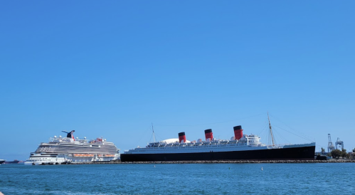 A modern cruise ship next to the historic Queen Mary ship, both docked in a calm harbor under a clear blue sky.