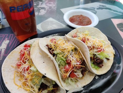 Three tacos filled with beef, lettuce, tomatoes, and cheese, served with a drink and a side of salsa.