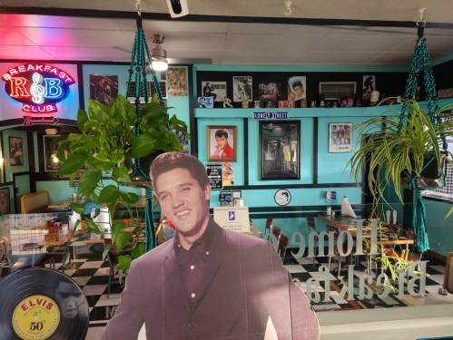A diner interior featuring a life-size Elvis cutout, vibrant decor, plants, and vintage music-themed artwork.