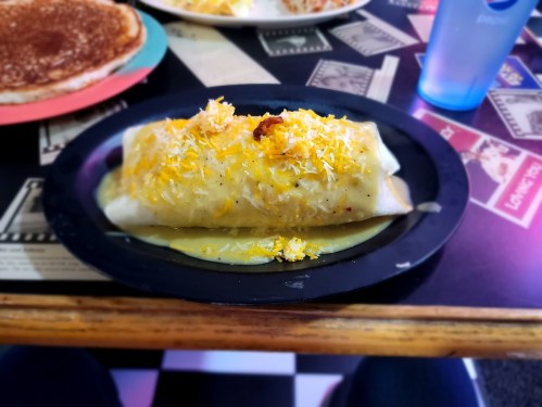 A plate with a large burrito topped with cheese and sauce, served alongside a drink on a patterned table.